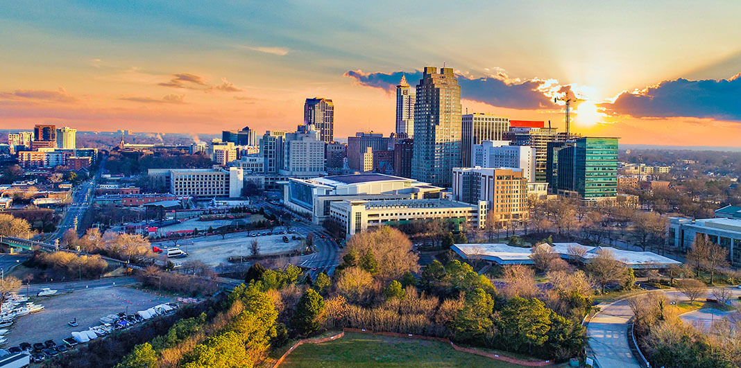 A city skyline with tall buildings and construction cranes at sunset, with trees and parking lots in the foreground.