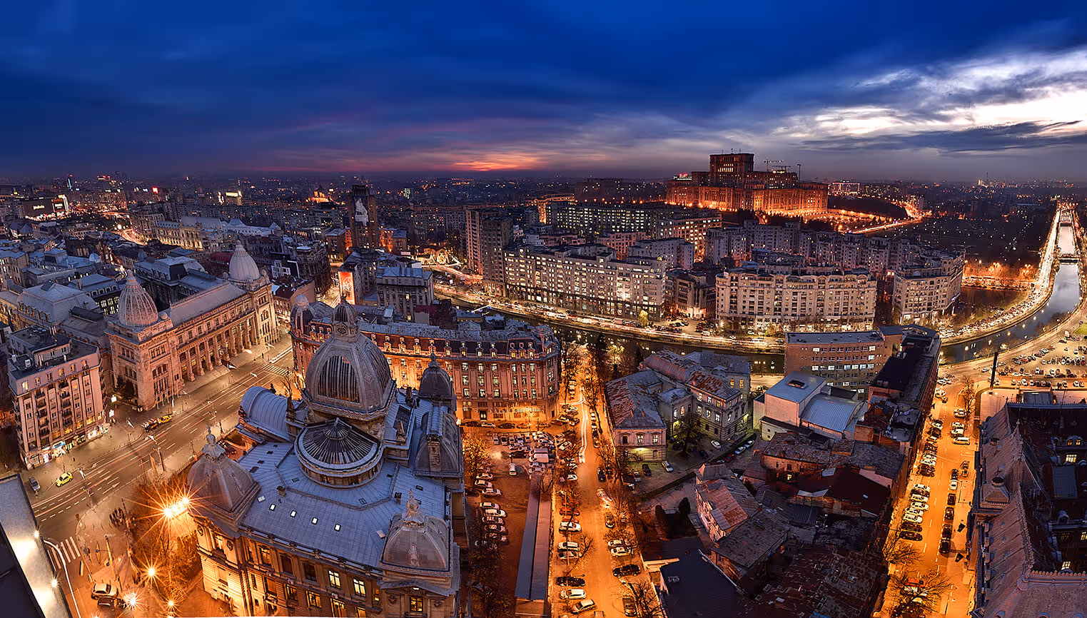 Aerial view of a cityscape at dusk with illuminated streets, domed buildings, and a river on the right side.