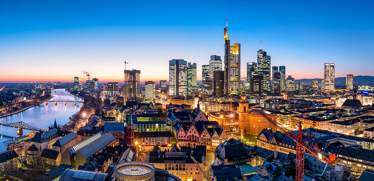 Cityscape at dusk with illuminated skyscrapers, river, bridges, and a mix of modern and historic buildings.