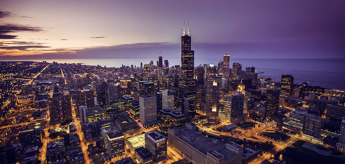 Aerial view of a city skyline with illuminated buildings at dusk, under a purple and orange sky.