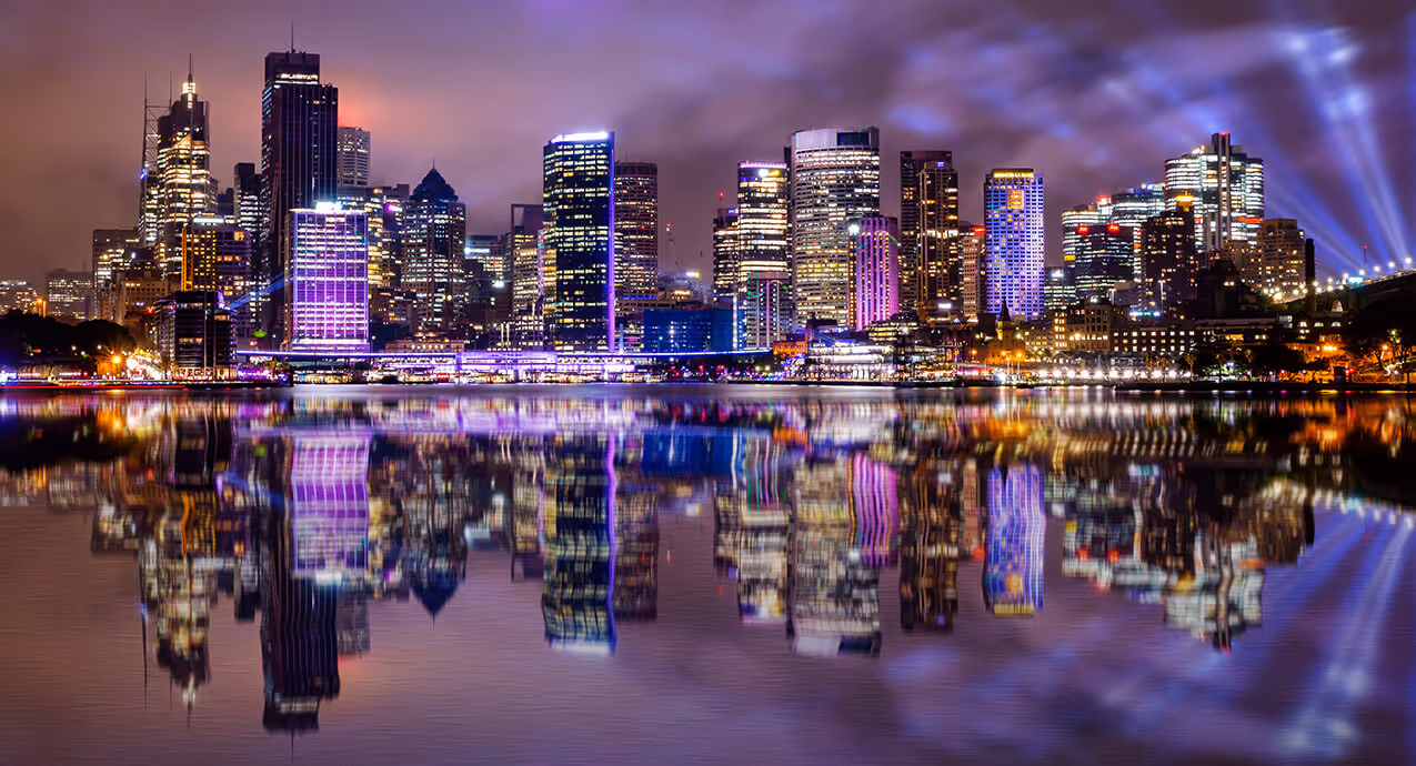 A city skyline at night with numerous illuminated buildings reflected in calm water.