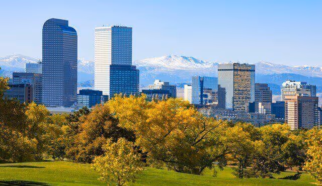 Tall modern buildings rise above a park with green trees and distant snow-capped mountains under a clear blue sky.