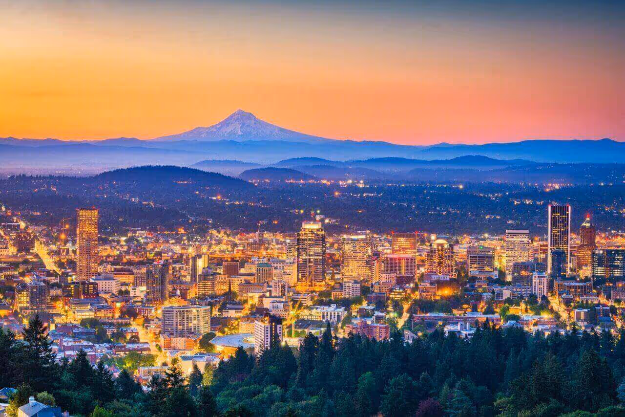 Cityscape at dusk with illuminated buildings in the foreground and a snow-capped mountain in the distance under a colorful sky.
