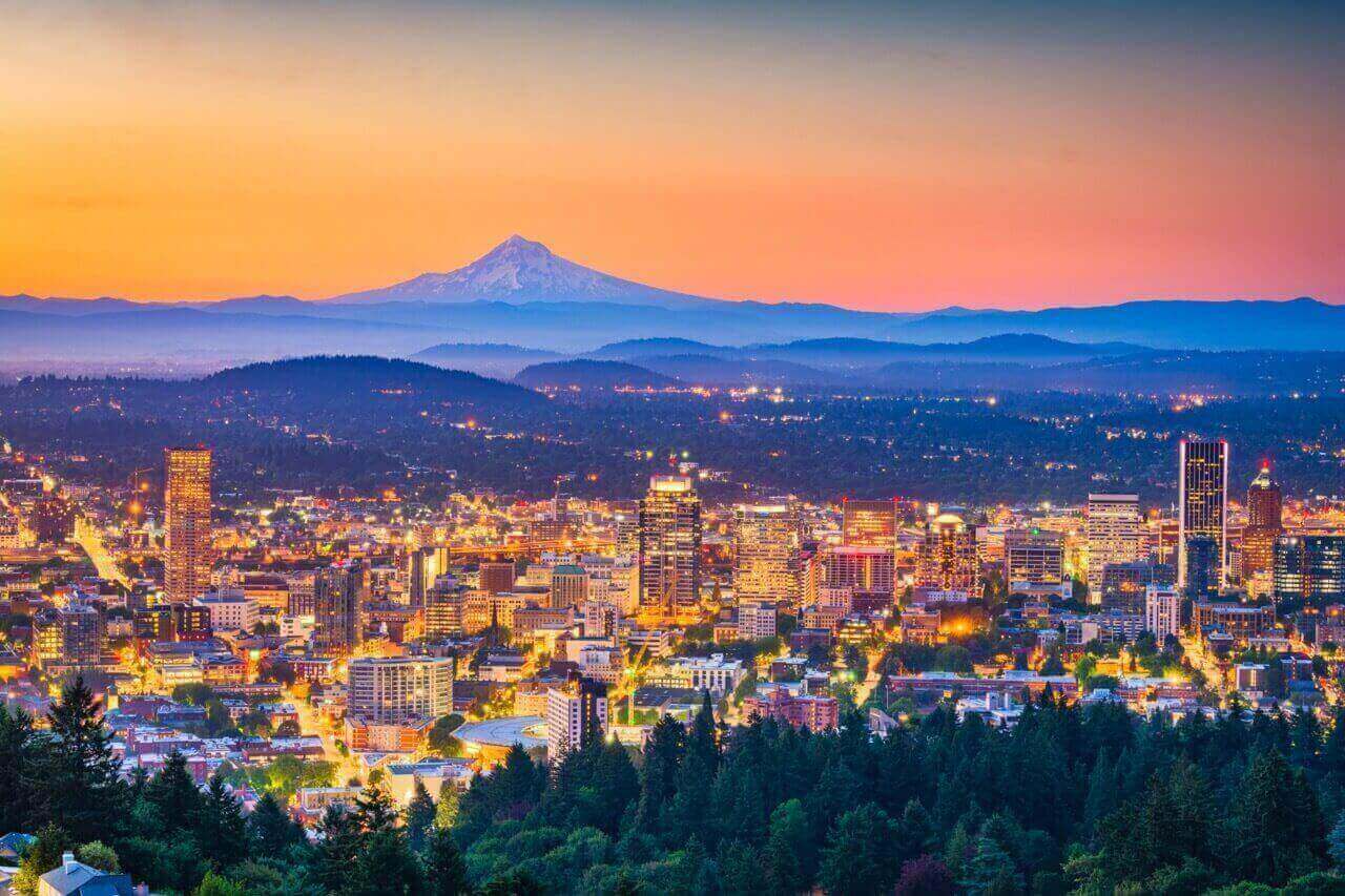 Cityscape at dusk with illuminated buildings in the foreground and a snow-capped mountain in the distance under a colorful sky.