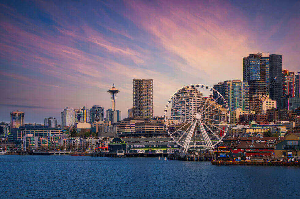 A waterfront cityscape with a large Ferris wheel, tall buildings, and a tower under a colorful sky at sunset.