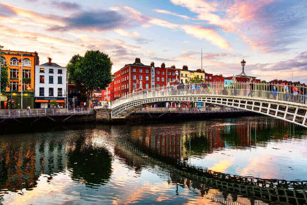 A curved pedestrian bridge crosses a river, with colorful buildings and trees lining the waterfront at sunset.