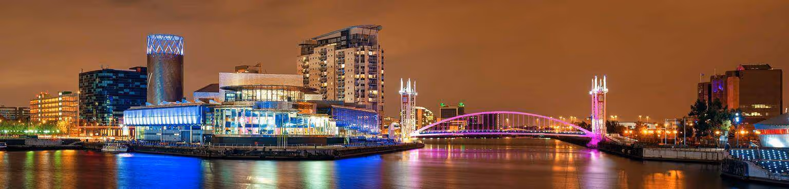 A nighttime cityscape with modern buildings, colorful lights, and a bridge reflecting on the water.