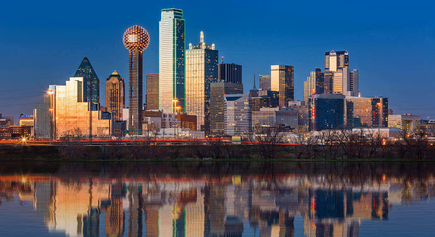 A city skyline at dusk with tall buildings illuminated and their reflections visible in water in the foreground.