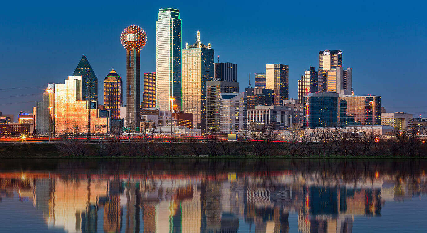 A city skyline at dusk with tall buildings illuminated and their reflections visible in water in the foreground.