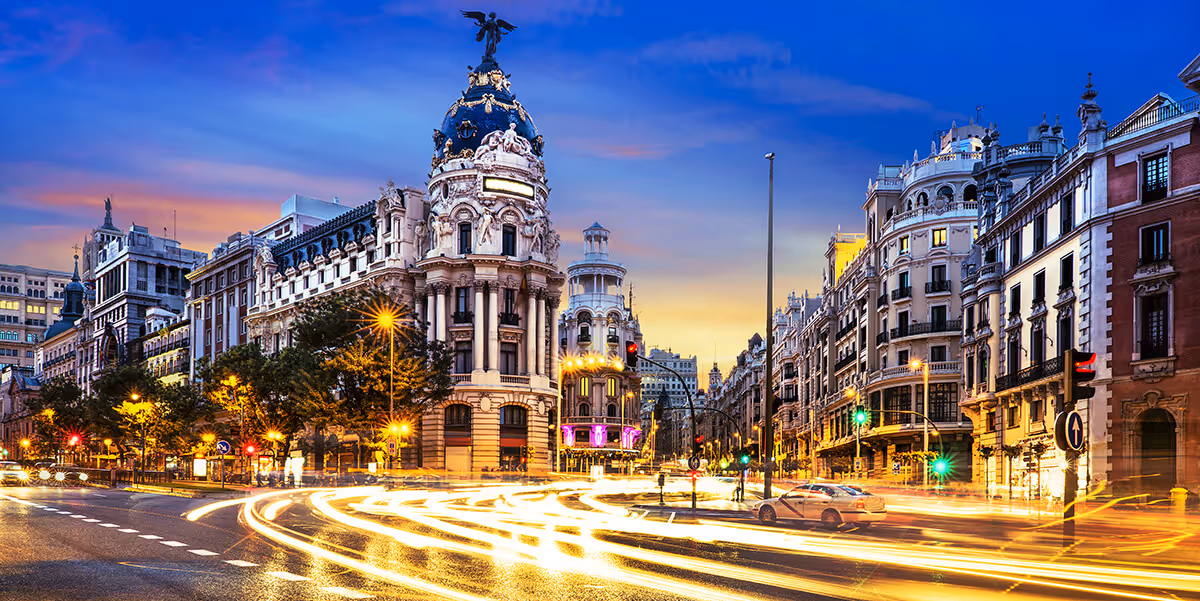 A city street scene at dusk with ornate historic buildings, moving cars, and light trails from traffic.