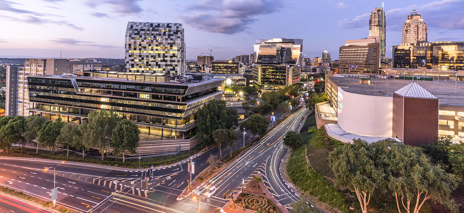 Modern cityscape at dusk with multiple office buildings, tree-lined streets, and car light trails on the roads.