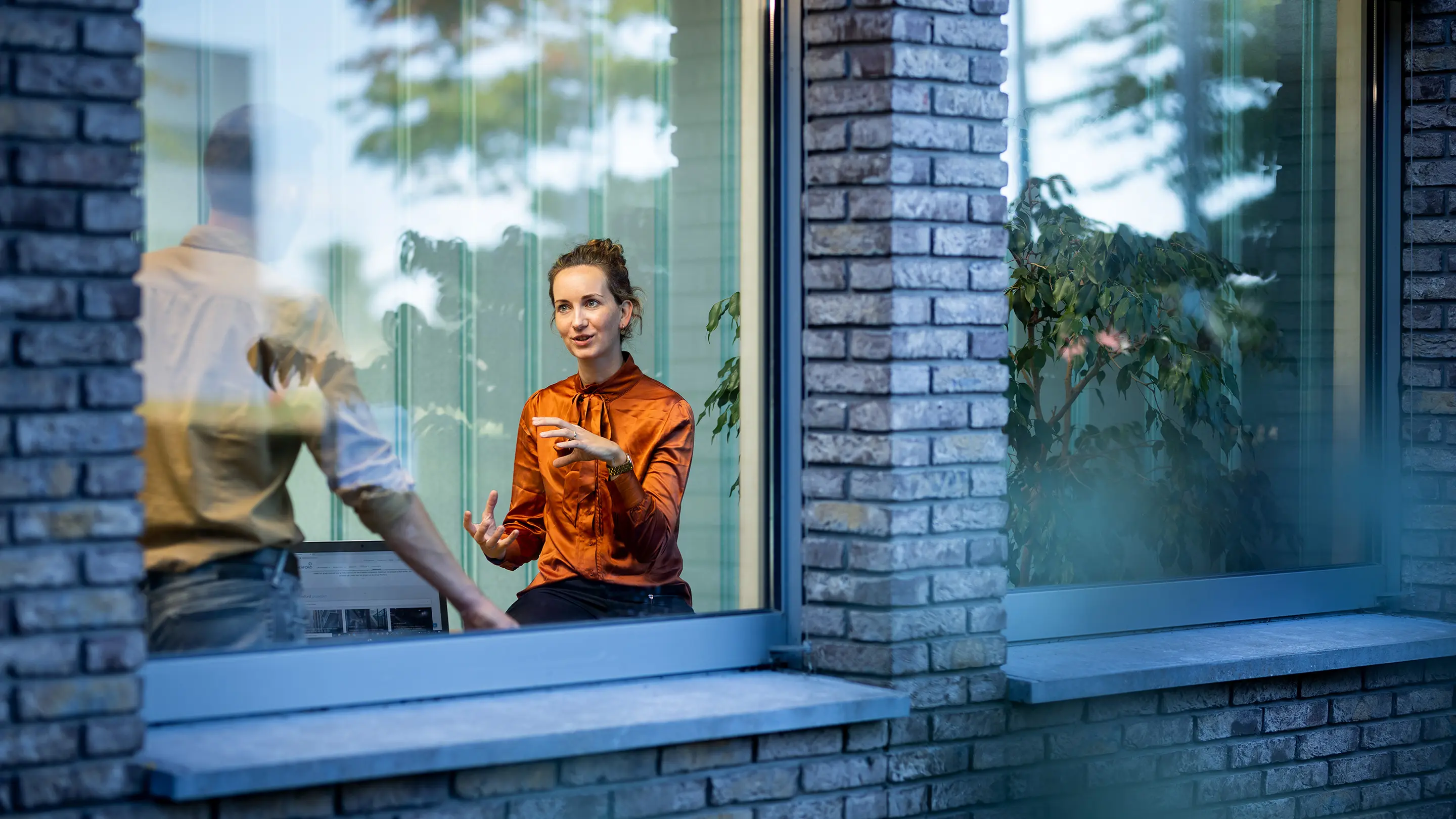 Two colleagues in conversation inside an office, viewed through a large window with reflections of trees.