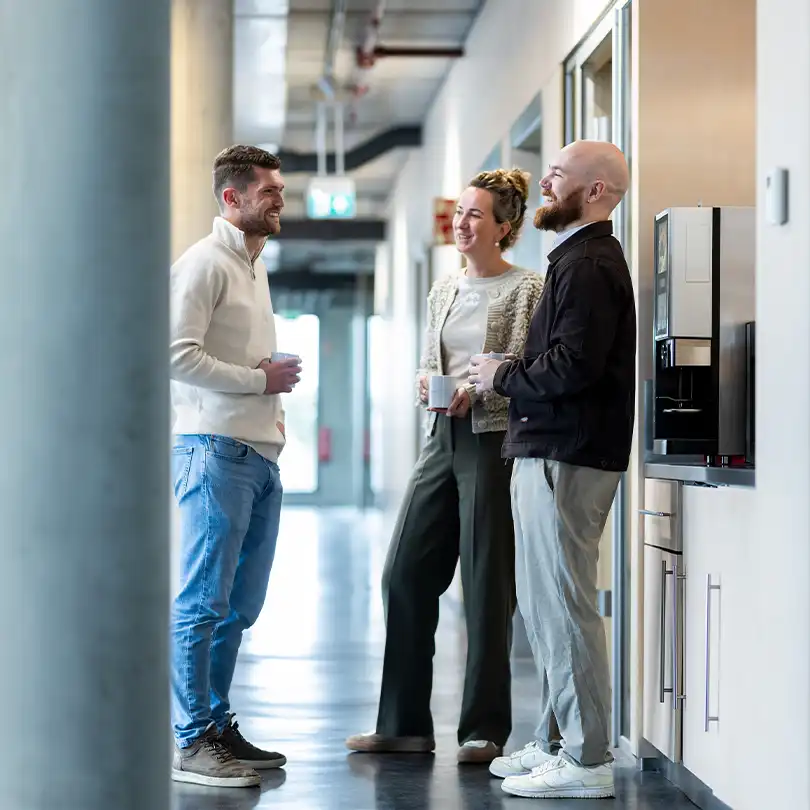 Three Merford office colleagues chatting and laughing together during a coffee break in the hallway.