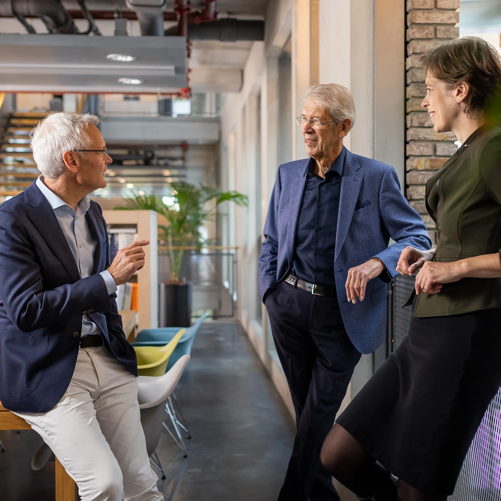 Three colleagues in professional attire having a friendly conversation in a modern office hallway.