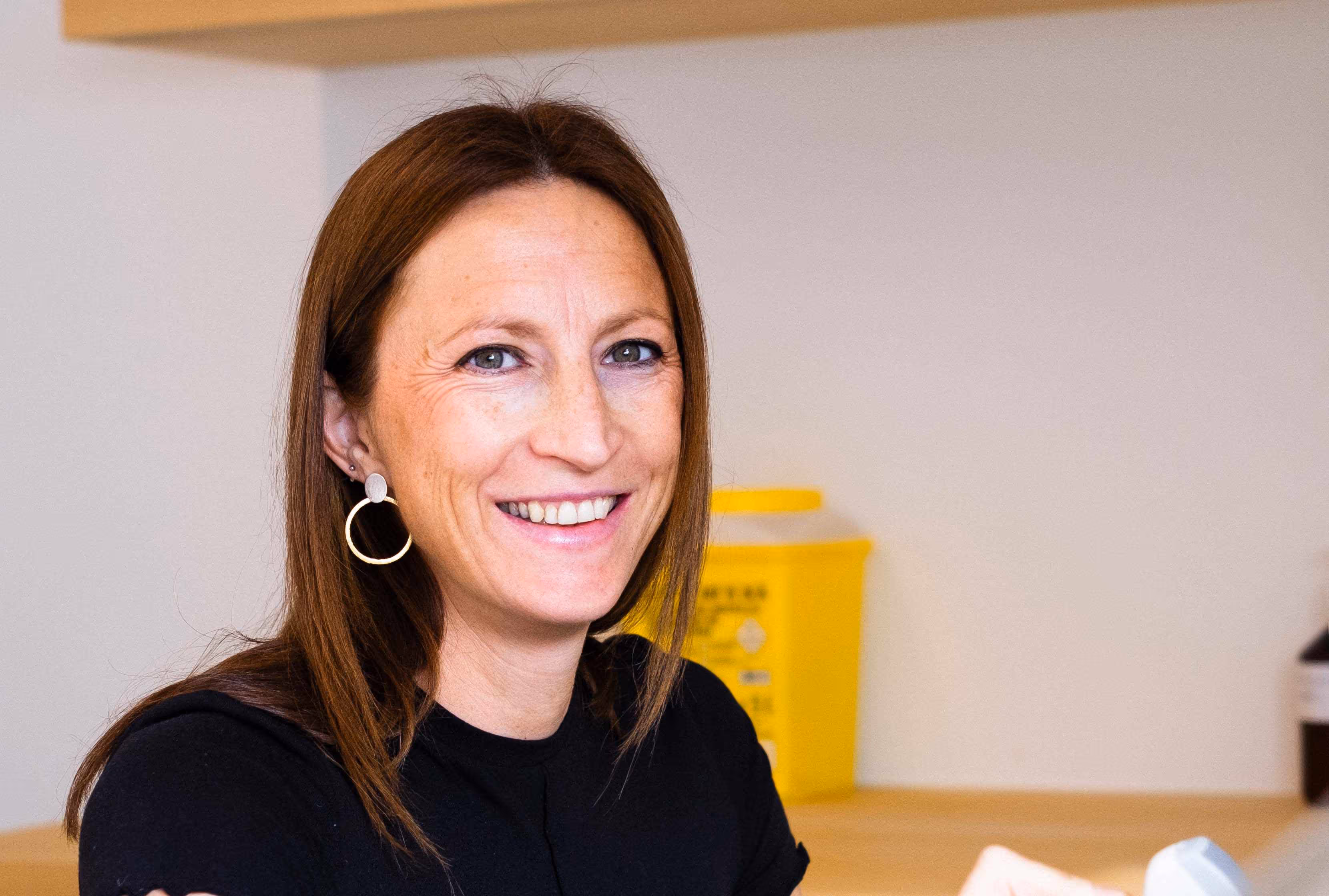 Vrouw met lange bruine haren en grote oorbellen lacht terwijl ze naar de camera kijkt.