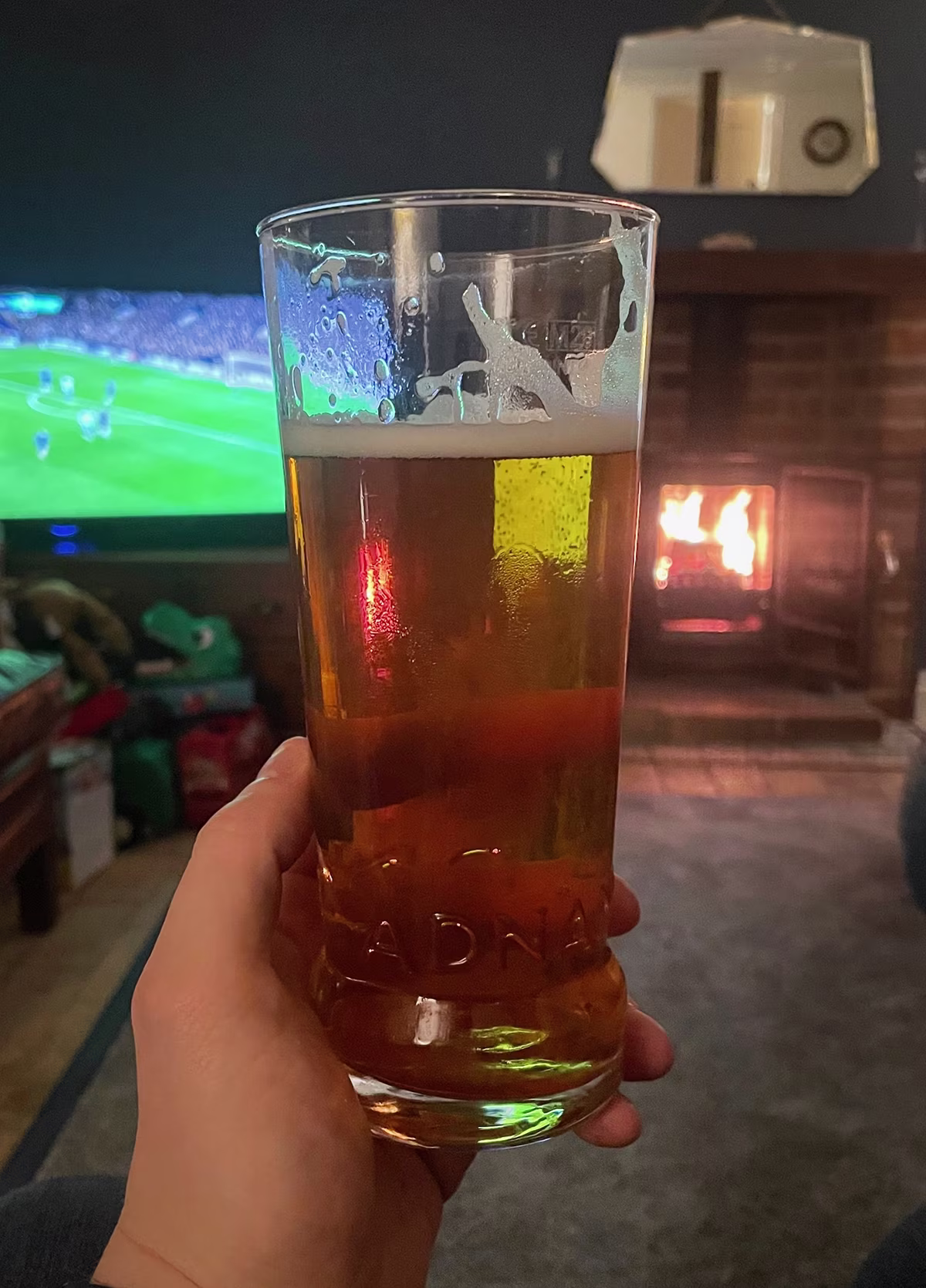 Hand holding a pint of beer in a cozy room with a lit fireplace and a football game on TV in the background.