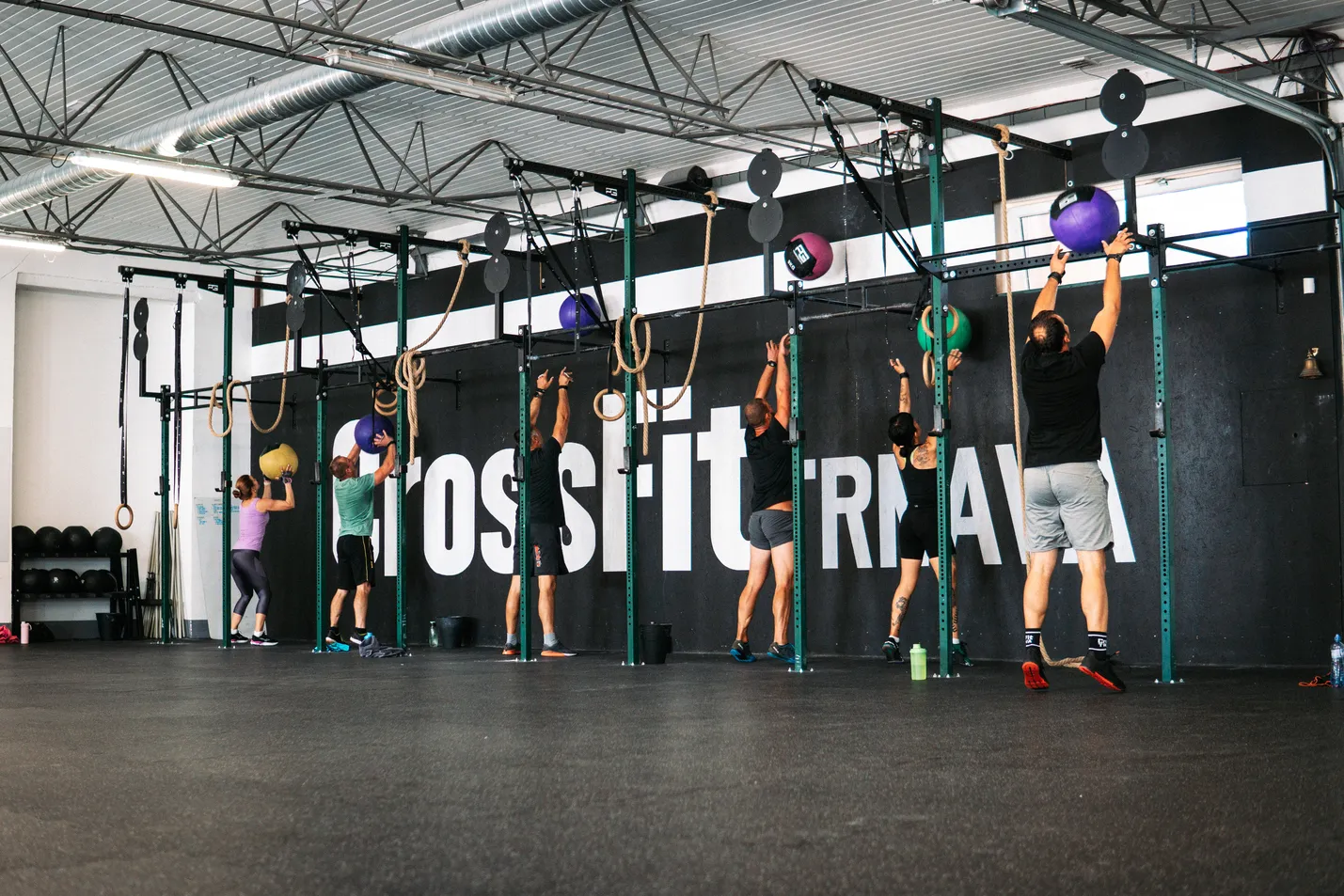 Skupina ľudí trénuje v CrossFit telocvični, hádžu medicine ball o stenu.