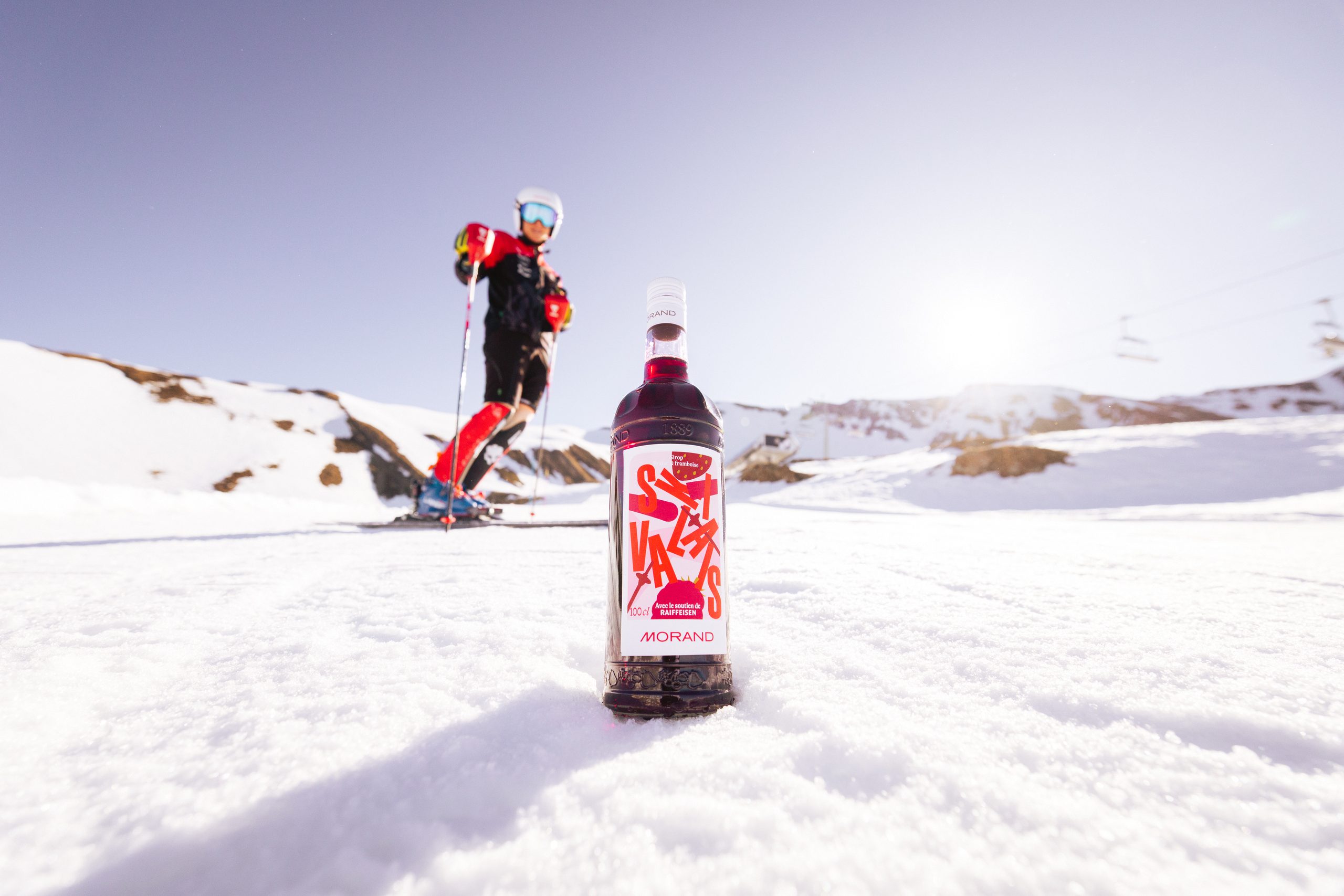 Bouteille Morand aux couleurs de Ski Valais posée dans la neige, un skieur en arrière-plan
