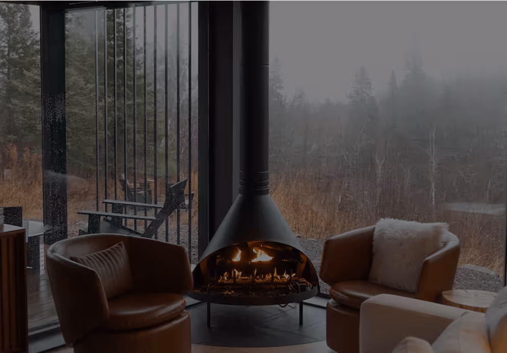 Coin salon avec cheminée suspendue allumée entre deux fauteuils en cuir devant une baie vitrée donnant sur un paysage automnal brumeux.