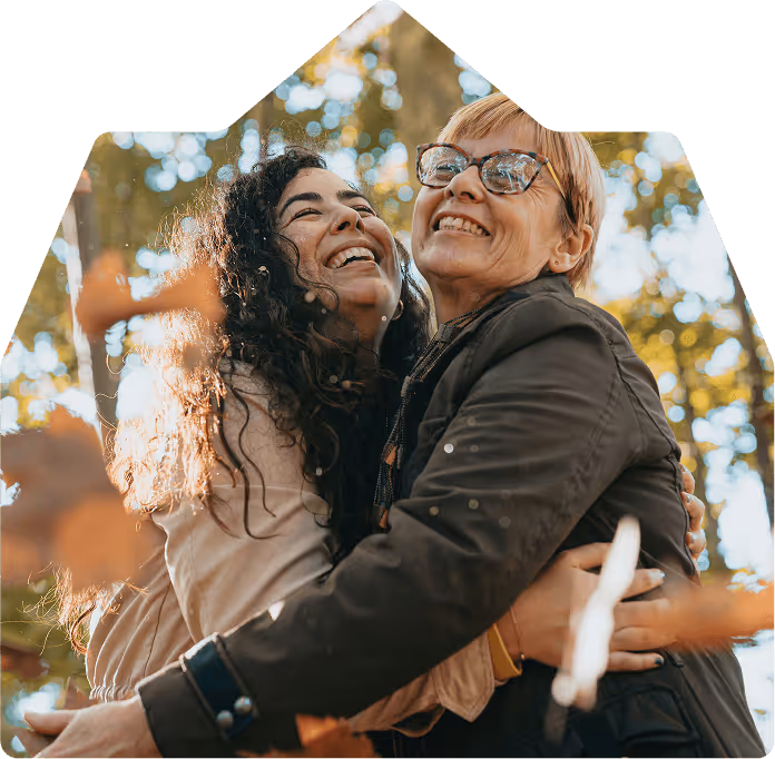 Two women happily embracing outdoors with autumn leaves and trees in the background.