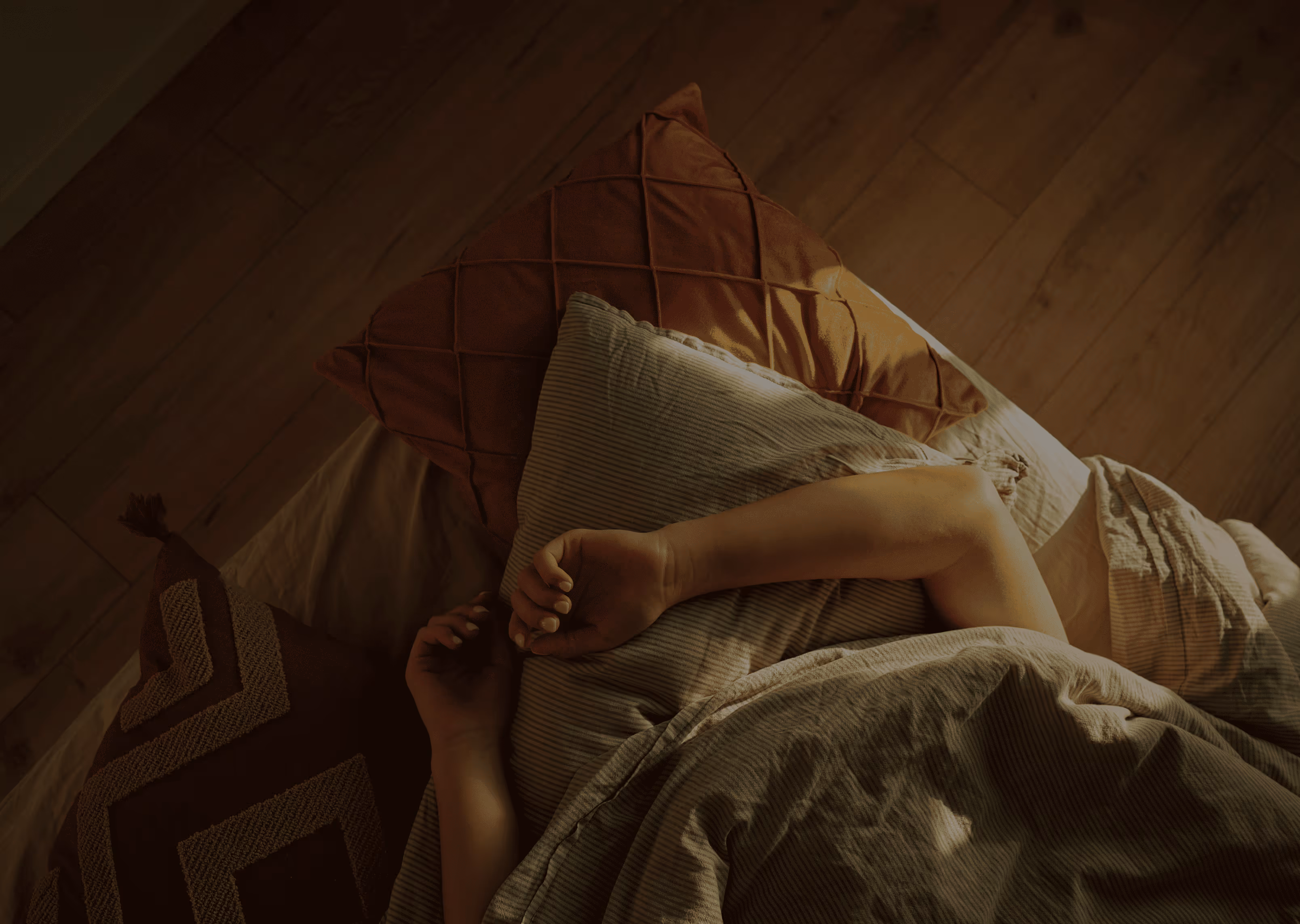 Person sleeping with arms resting on pillows and covered by a blanket on a wooden floor.