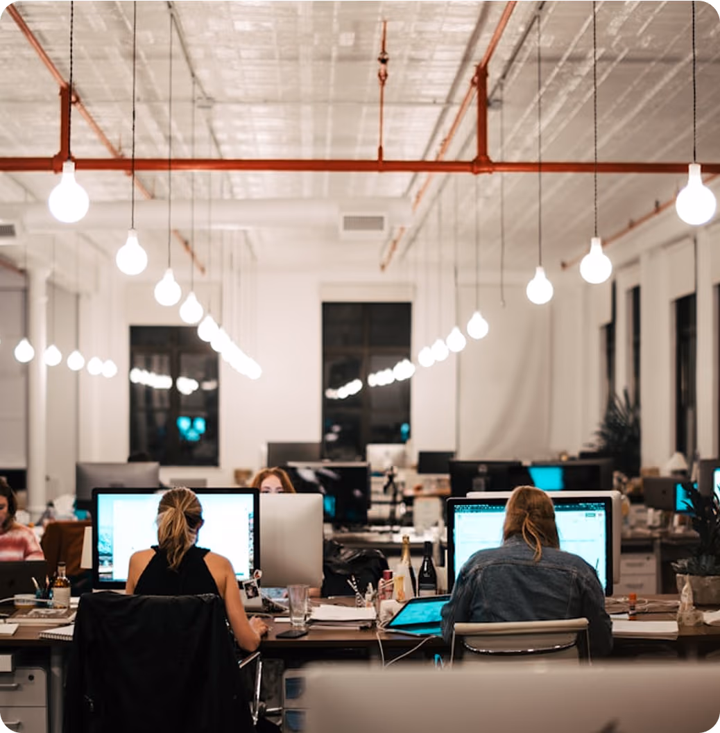 Vue arrière de plusieurs personnes travaillant sur des ordinateurs dans un bureau moderne éclairé par des ampoules suspendues.