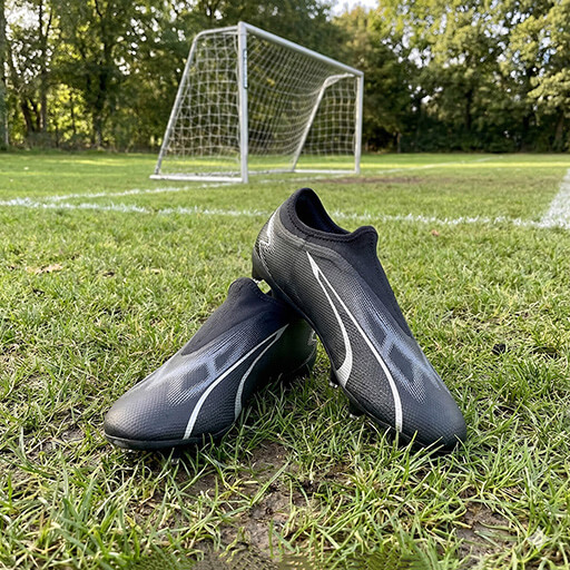 Puma Ultra Match Jr black kids’ football boots displayed on a grass field with a goal behind them, showing their sleek laceless design and silver detailing