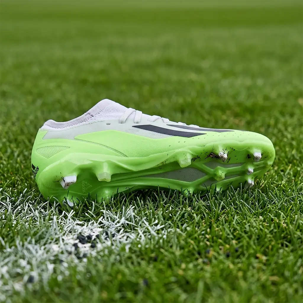 A detailed look at the lime green and white Adidas X Crazyfast .3 FG Junior football boot, showing the stud pattern and outsole while resting on a natural grass pitch. This authentic field-test shot highlights the traction and stability features for junior players