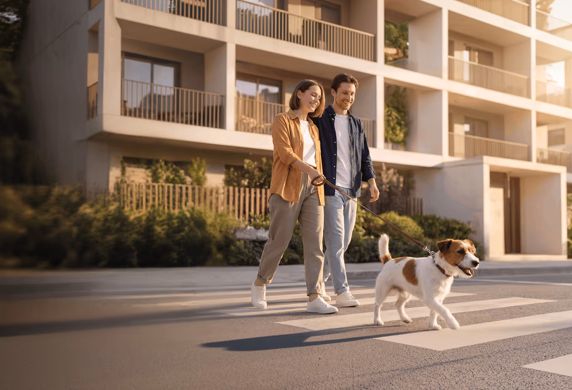 A smiling couple walking a dog on a leash across a street in front of an apartment building.