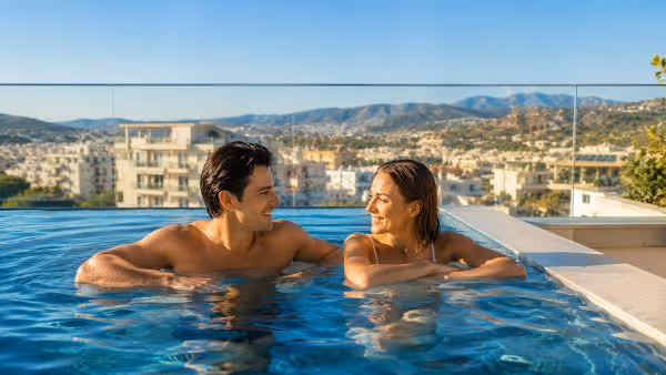 Man and woman smiling at each other in an outdoor pool with city and mountain view.
