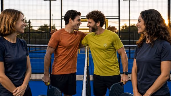 Two men smiling and shaking hands at a net on a paddle tennis court with two women watching.