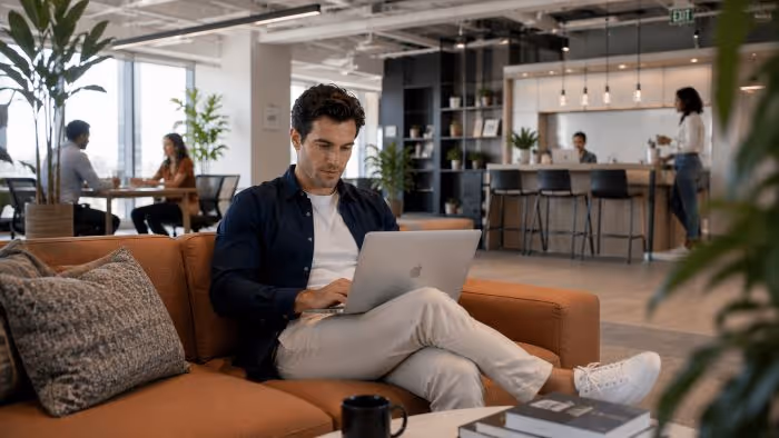 Man sitting on an orange sofa using a laptop in a modern open office lounge area.