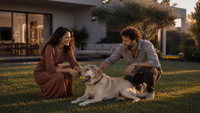 A smiling couple kneels on grass petting a large yellow Labrador resting with a ball in its mouth.
