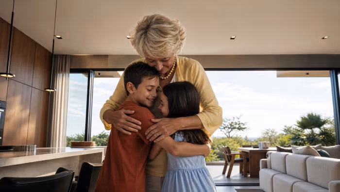 Smiling elderly woman hugging two children inside modern living room near large windows.