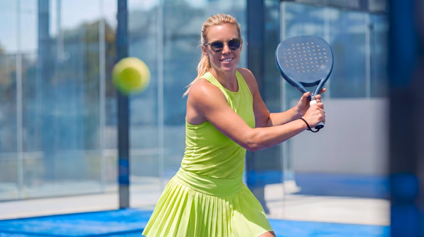 Woman in yellow sports outfit playing paddle tennis on glass court hitting ball with paddle.