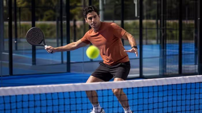 Man in orange shirt playing paddle tennis, hitting yellow ball on blue court near net.