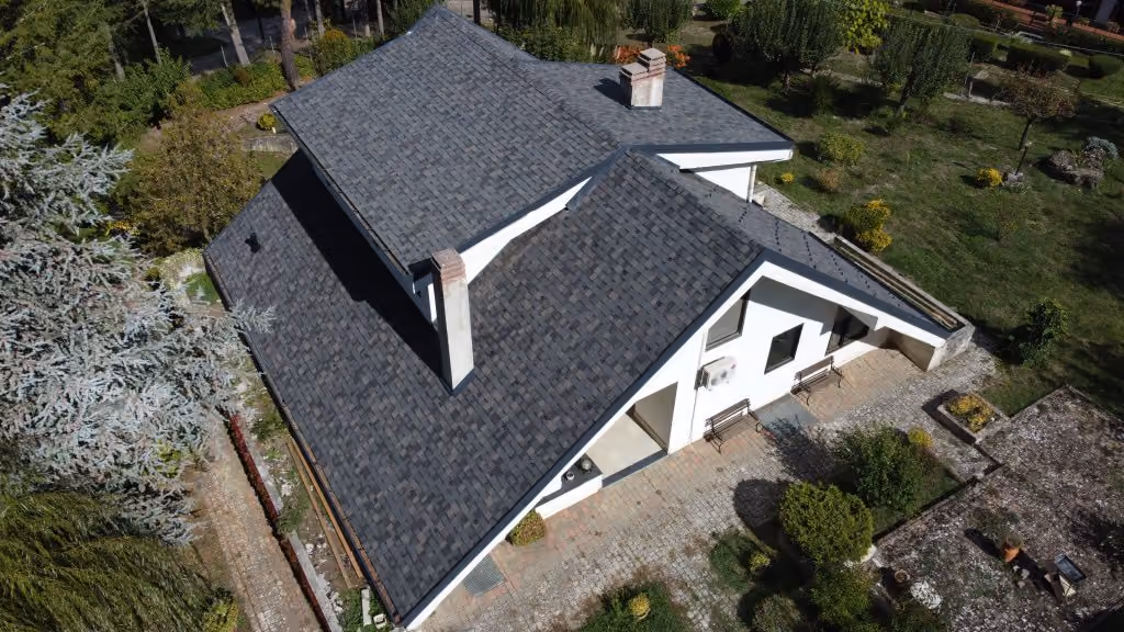 Aerial view of a white house with a dark gray complex pitched roof surrounded by green garden.