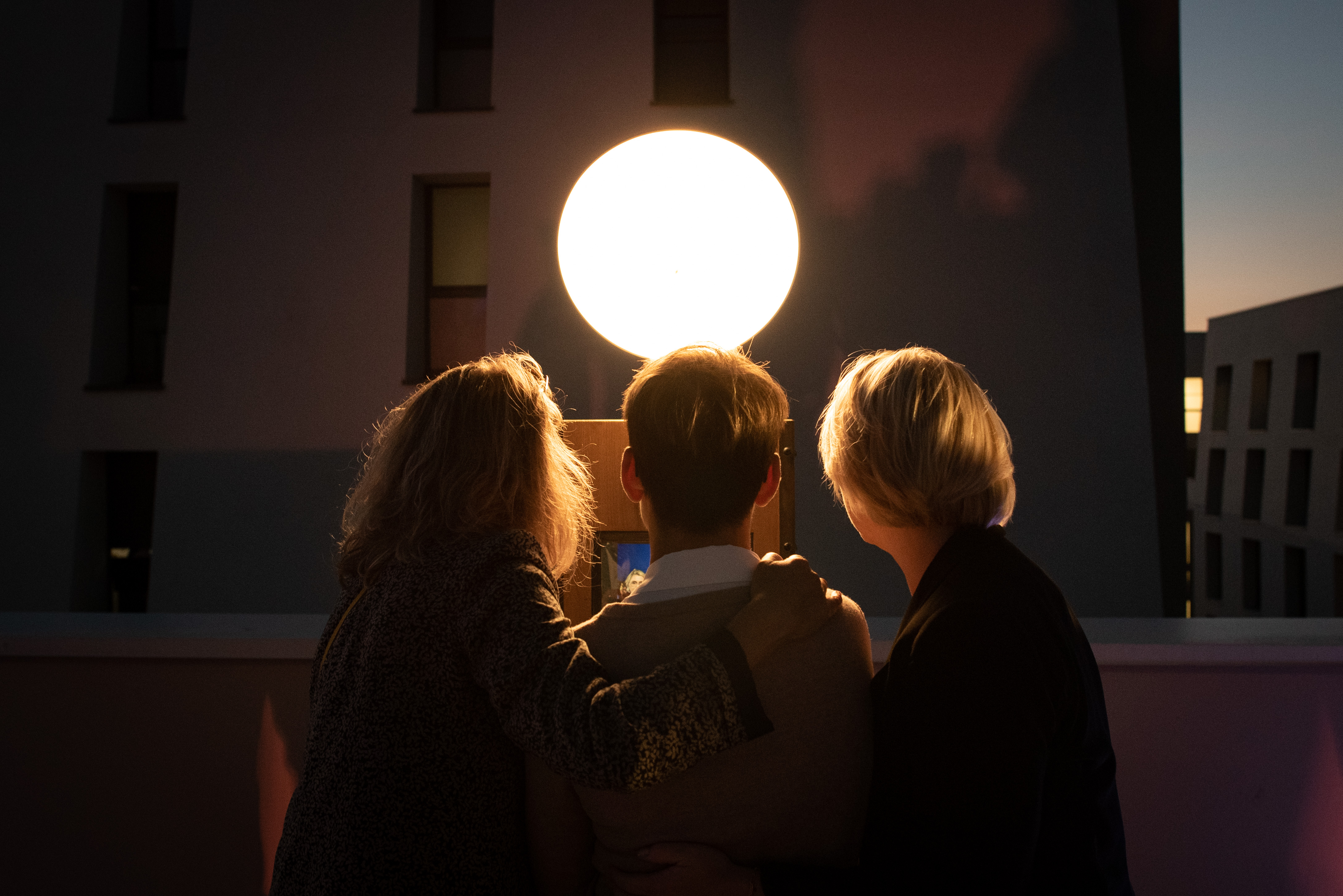 Trois personnes de dos regardant un écran éclairé par une grande sphère lumineuse dans une ambiance nocturne urbaine.