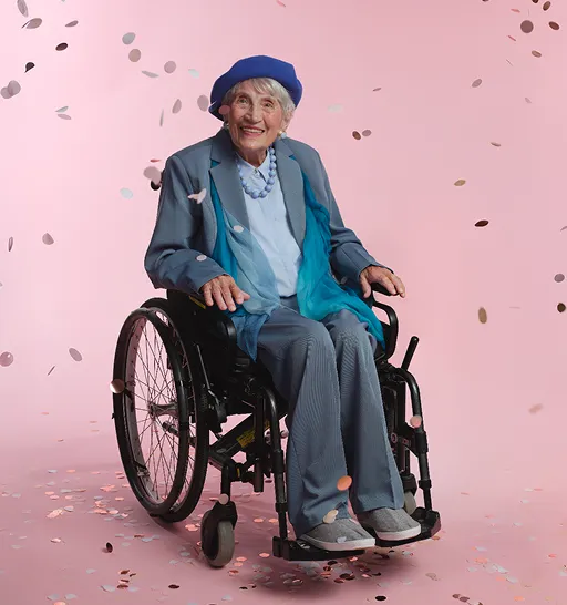 Eine charmante ältere Dame im Rollstuhl, elegant gekleidet. Um sie herum fliegen Konfetti.