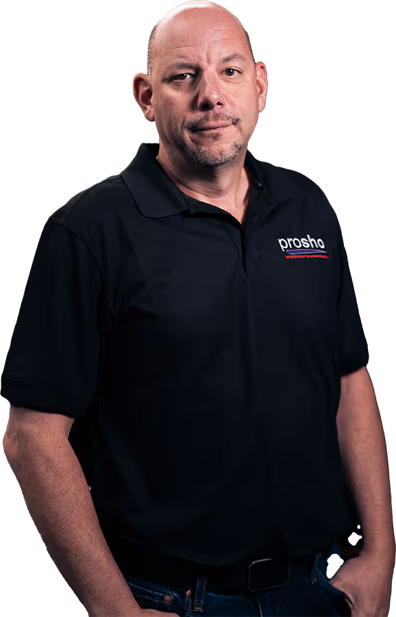 Bald man wearing a black Prosho polo shirt standing with one hand in his pocket against a dark blurred background.