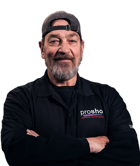 Middle-aged man with a gray beard wearing a backward black cap and a black shirt with 'prosho' logo, arms crossed and smiling.