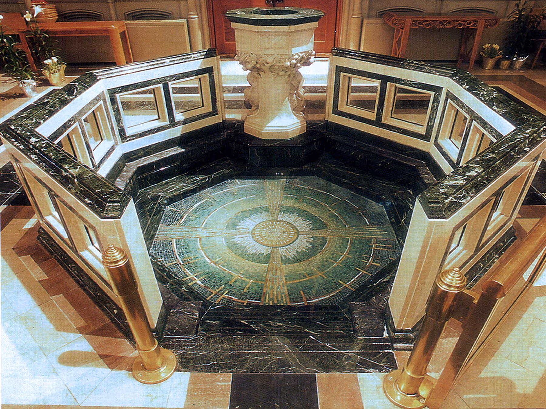 Ornate octagonal baptismal font with black and white marble surround and mosaic floor inside a church.