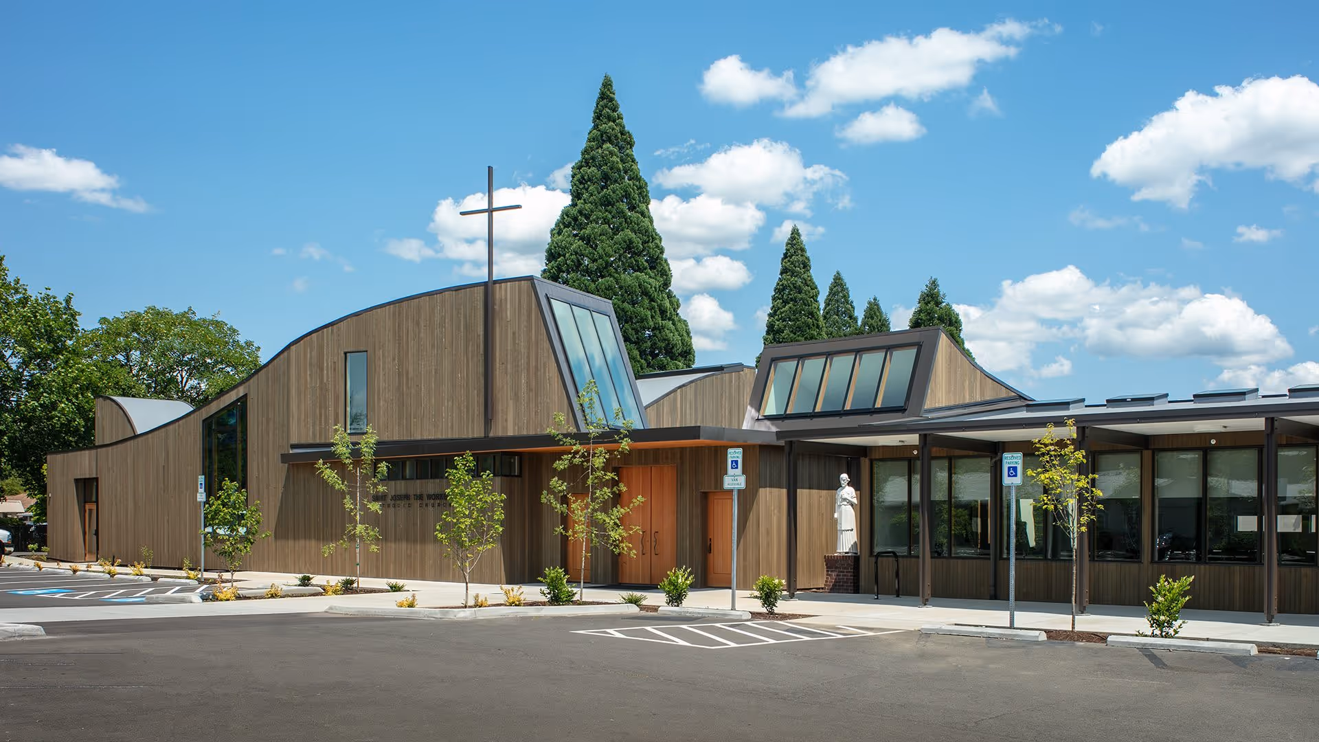 Modern wooden church building with large windows, a cross on the roof, and a statue near the entrance, under a blue sky with scattered clouds.