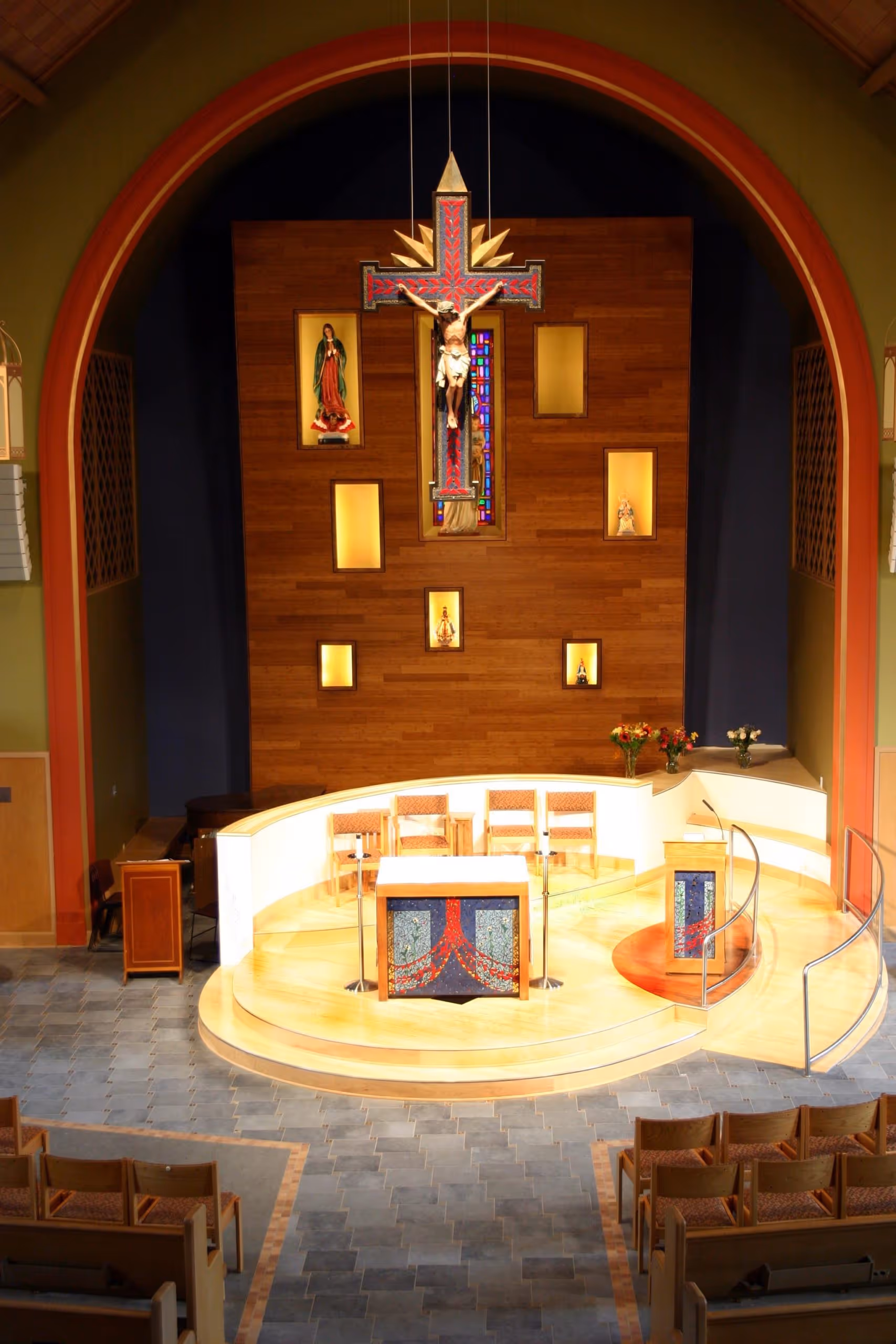 Interior of a church altar area with a crucifix hanging above, wooden backdrop with religious statues, curved platform with altar and chairs, and tiled floor.
