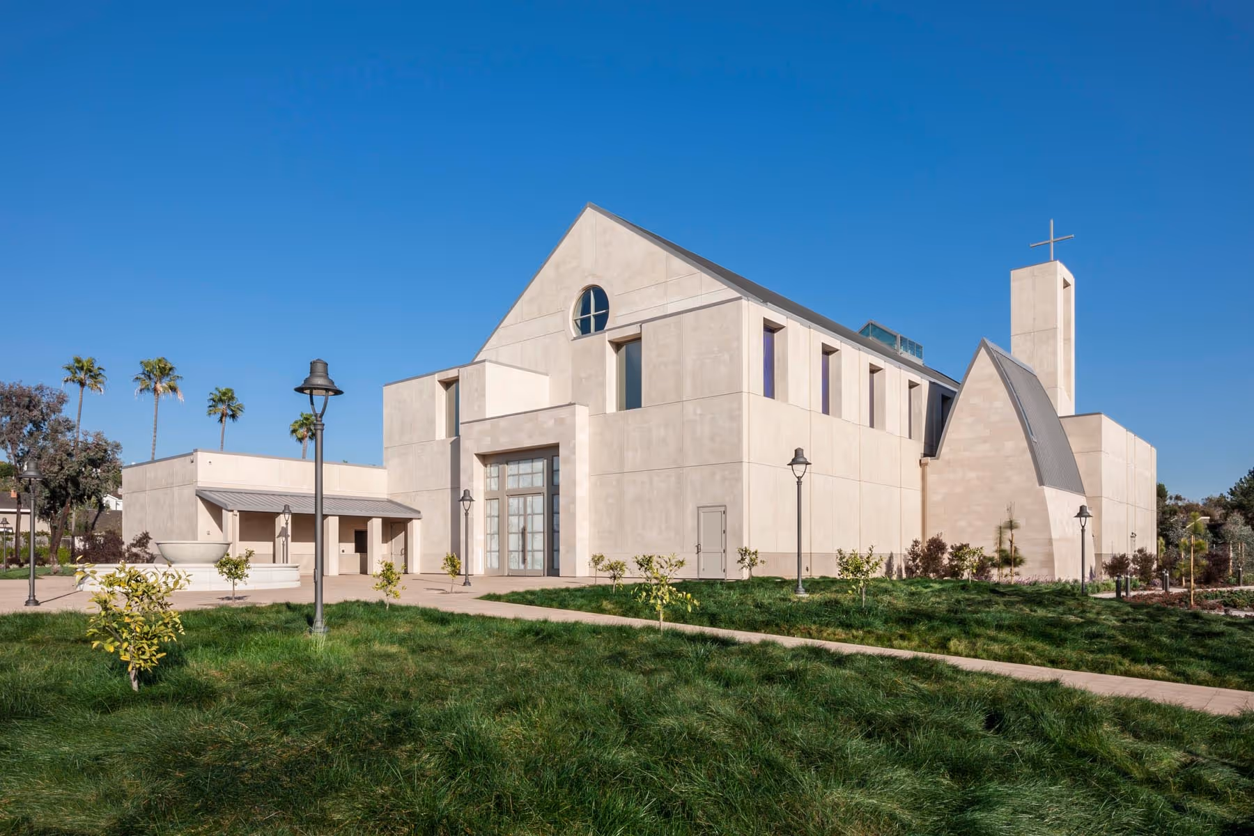 Modern beige church building with a tall tower topped by a cross, surrounded by green grass and lamp posts under a clear blue sky.