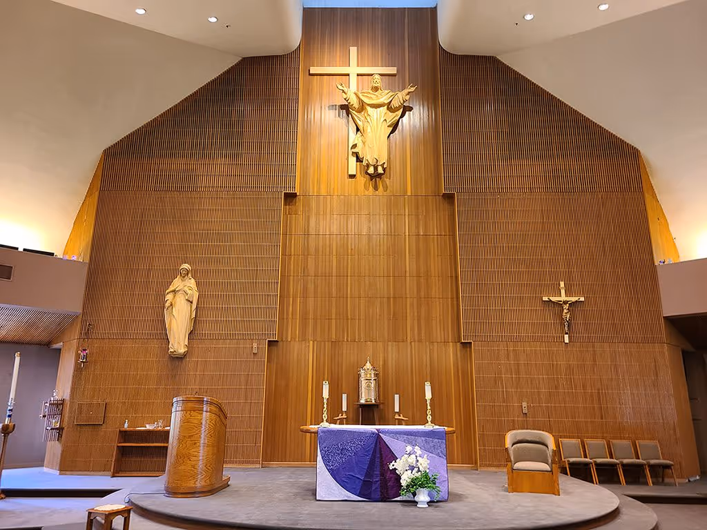 Interior view of a church altar with a wooden backdrop, a large statue of Jesus on a cross above, statues on either side, altar table with candles and flowers in front.