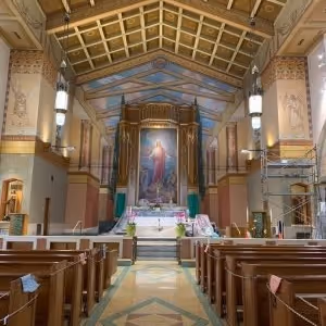 Interior view of a church with wooden pews, a tiled floor, ornate ceiling, and a large religious painting behind the altar.