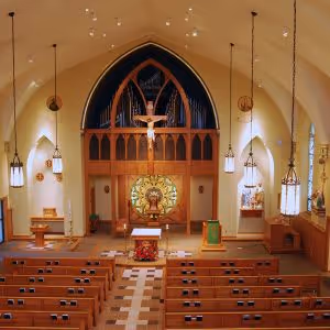 Interior view of a church with wooden pews, hanging pendant lights, a large crucifix, organ pipes, and an altar adorned with flowers and religious symbols.