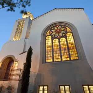 White building with a large colorful stained glass window and a tower illuminated at dusk.