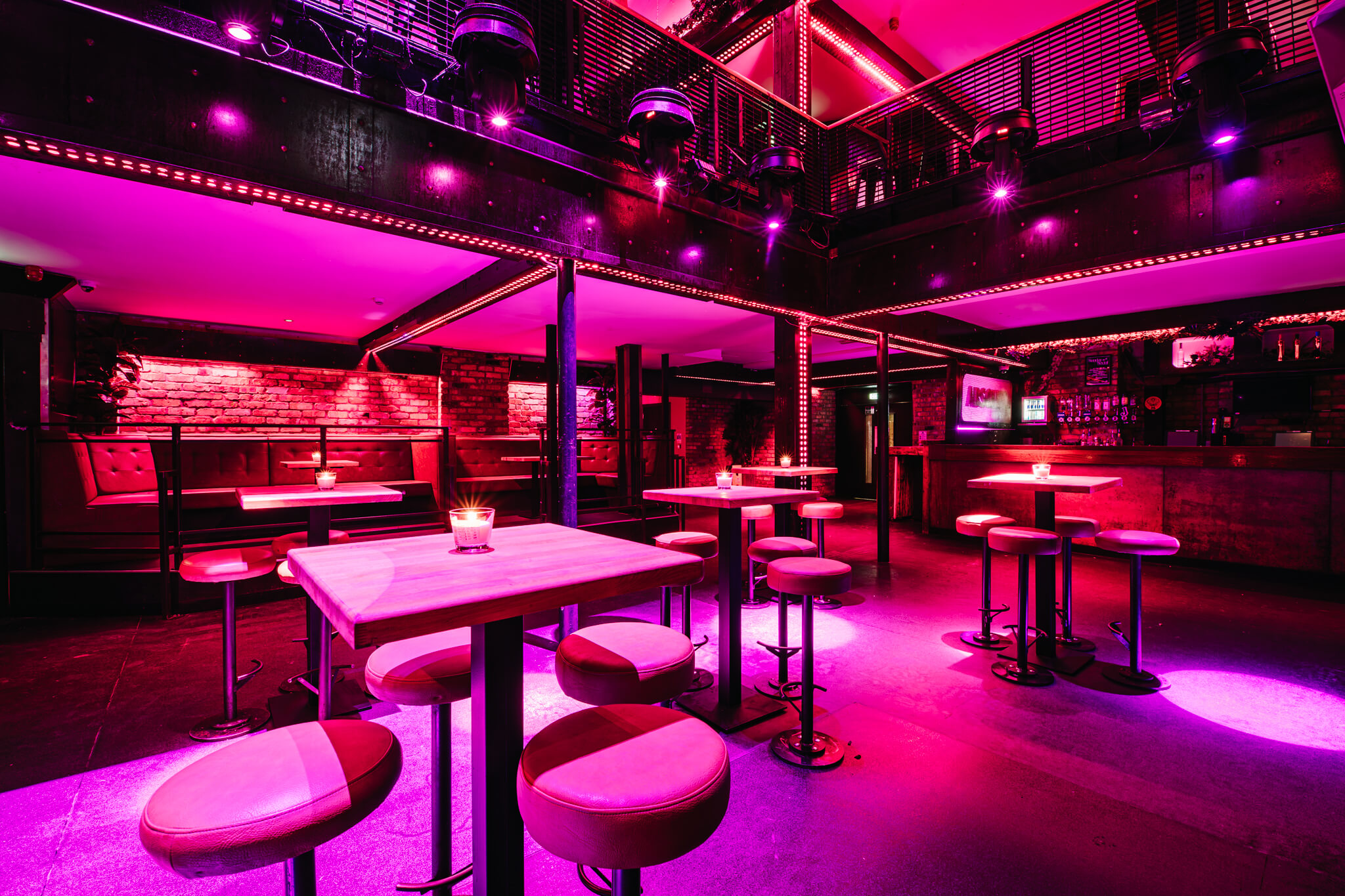 Interior of a nightclub showing tables and chairs under purple lighting. The space has a modern design with exposed brick walls and a bar in the background, suitable for events like parties or venue hire.
